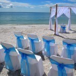 A beach wedding with white chairs and blue bows