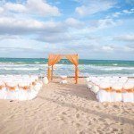 A beach wedding with white chairs and orange bows.