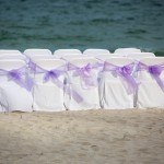 A row of white chairs with purple bows on the beach.