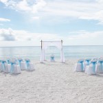 A beach wedding with white chairs and blue bows