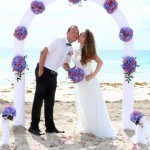 A newly married couple kissing under an arch on the beach.