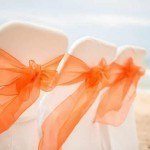Three chairs with orange bows on them at the beach