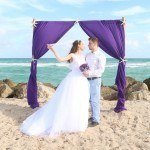 A bride and groom standing on the beach under an arch.