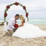 A bride and groom sitting on the beach under an arch.