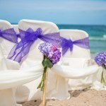 A row of white chairs with purple flowers on the beach.