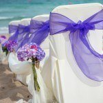 A row of white chairs with purple bows on the beach.