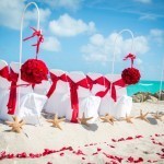 A beach wedding with red flowers and white chairs