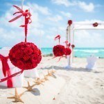 A beach wedding with red flowers and white decorations.