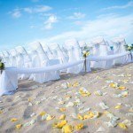 A beach wedding with white chairs and yellow flowers.