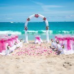 A beach wedding with pink flowers and white chairs.