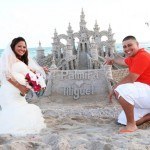 A couple poses for a picture in front of a sand castle.