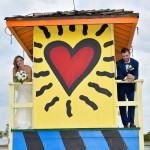 A bride and groom standing on the side of a yellow lifeguard tower.